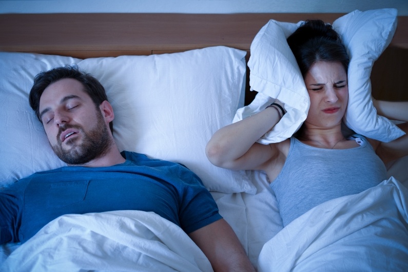 Frustrated woman covering her ears with pillow in bed next to man sleeping with mouth open