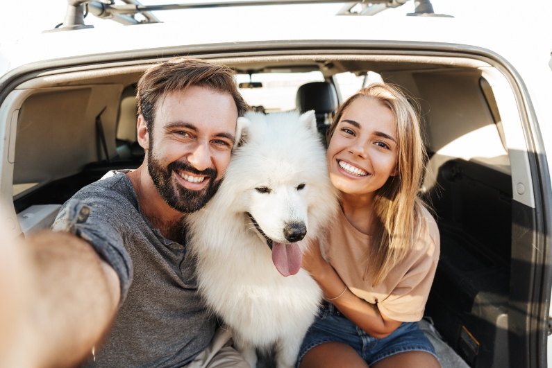 Man and woman smiling with fluffy white dog in trunk of car