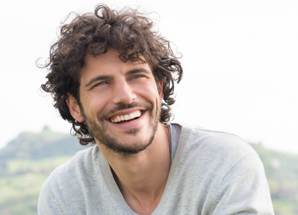 Young man with curly brown hair grinning outdoors on cloudy day