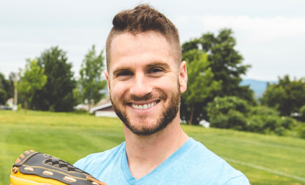 Man grinning outdoors with green field and trees in background