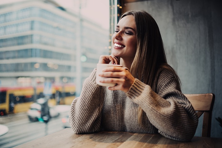 Woman in brown sweater in café holding coffee cup smiling gazing out the window